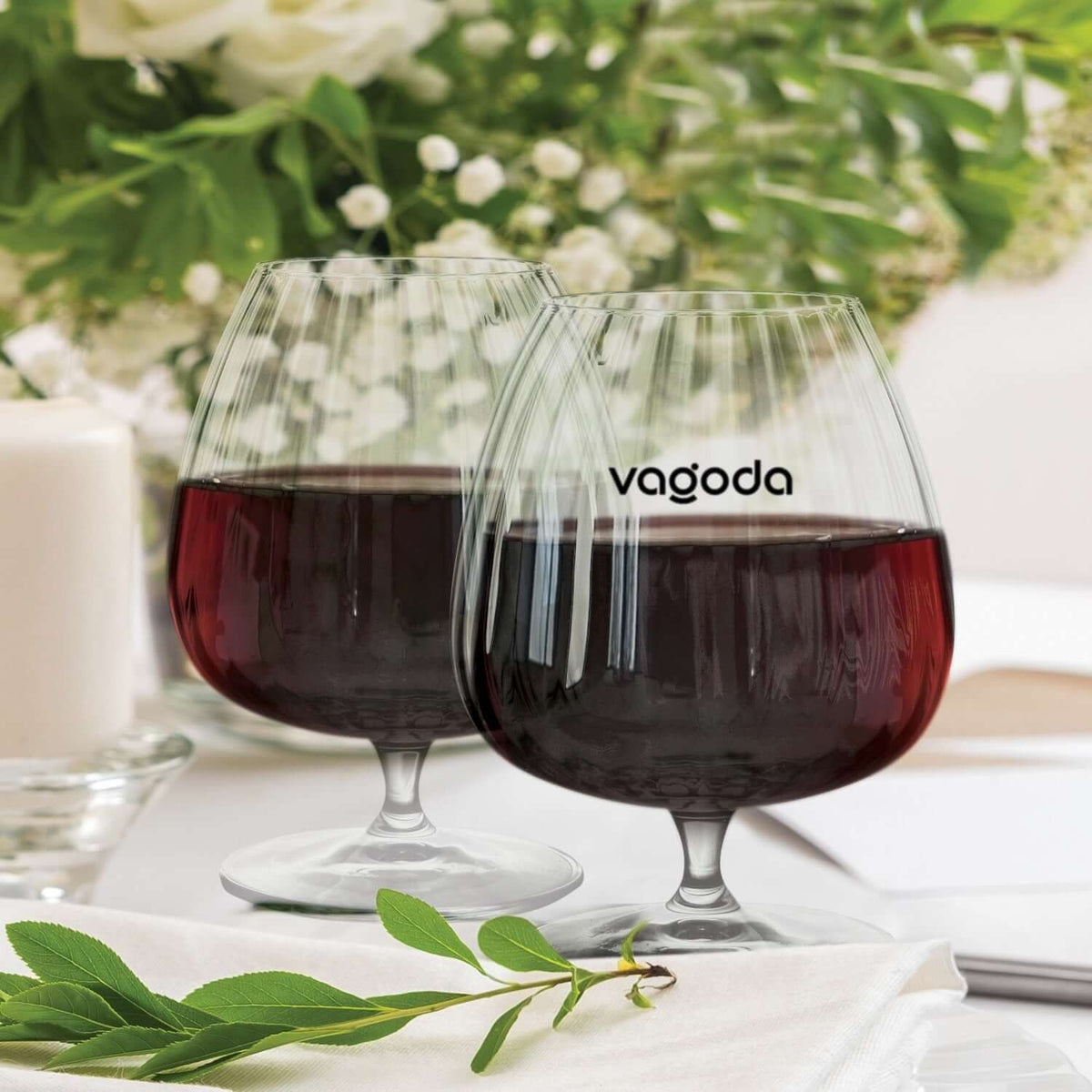 Two Vagoda red wine glasses filled with wine, surrounded by elegant decor and greenery.